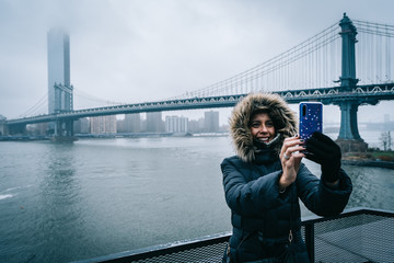 Tourist woman with warm clothes taking a selfie with the Manhattan Bridge on the background in...