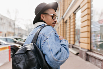 Outdoor photo from back of confident african man in denim jacket walking on the street. Stylish black guy looking at showcase.