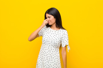 young pretty latin woman feeling disgusted, holding nose to avoid smelling a foul and unpleasant stench against flat wall - obrazy, fototapety, plakaty