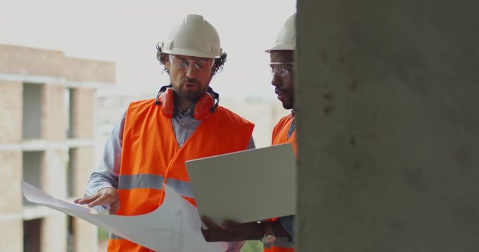 Two multiethnic men, architect with laptop computer and foreman with drafts and plans standing at the terrace of the building site of high building and talking over the constructing process.