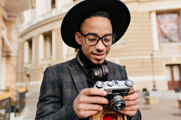 Curious african man in hat looking at camera. Outdoor portrait of black male photographer standing near beautiful building.