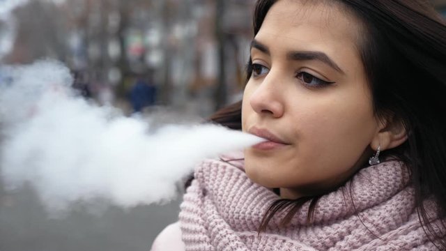 Portrait of brunette young woman relaxing on the street with vape-device. Vaping girl outdoors, slow motion