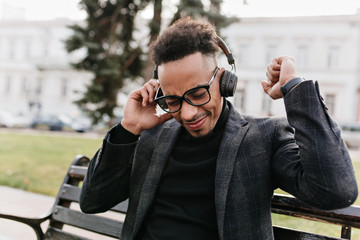 Pleased dark-haired african man wears black clothes chilling outdoor. Photo of relaxed guy in glasses enjoying music in headphones.