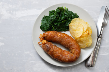 fried smoked sausage with potato chips and greens on white plate on ceramic background