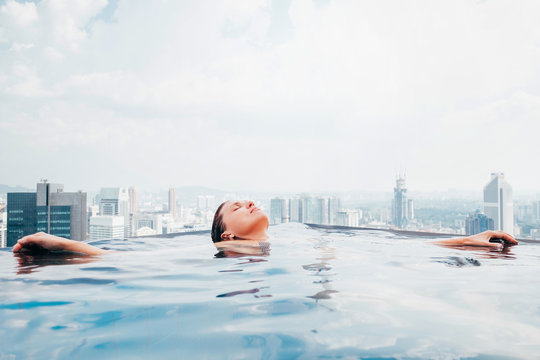 Young female relaxing in top roof infinity swimming pool with city view to the modern business center downtown with a skyscrapers and towers. Luxury vacation concept image.