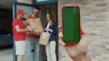Food delivery services. View of young man using smartphone in front of young cheerful couple receiving food from courier. Male hand with greenscreen chroma key mobile phone.