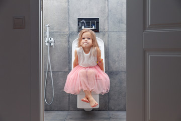 Cute little girl in white pink dress sitting on toilet with toilet paper on background of walls with gray tiles, view from open door.