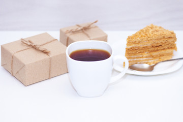 Slice of homemade layers honey cake, cup of coffe, present box or gift on wooden white background. Happy morning, copy space.