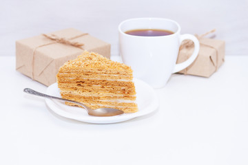 Slice of homemade layers honey cake, cup of coffe, present box or gift on wooden white background. Happy morning, copy space.