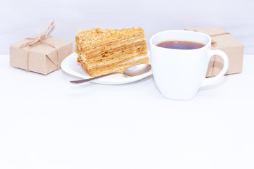 Slice of homemade layers honey cake, cup of coffe, present box or gift on wooden white background. Happy morning, copy space.
