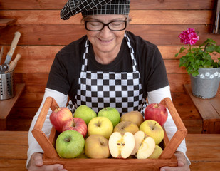 High angle view of an attractive and smiling senior woman holding a basket full of apples of various qualities and colors. Rustic background in recycled wood. One people only