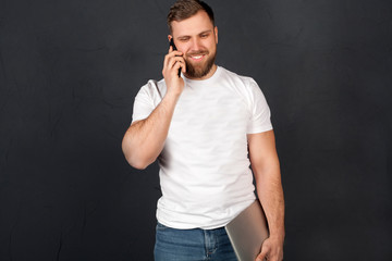 A man is talking on the phone holding a laptop in his hands, in a white t-shirt and blue jeans, on a gray background, copy space. The concept of business and new technology. 