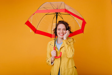 Gorgeous pale girl in autumn coat smiling with eyes closed under parasol. Studio portrait of stylish caucasian woman with wavy hair holding red umbrella.