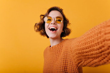 Laughing magnificent woman in funny yellow sunglasses making selfie. Studio portrait of relaxed...