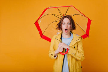Surprised brunette girl wears trendy bright coat posing with mouth open. Indoor photo of shocked...