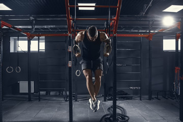 Obraz na płótnie Canvas Flying hard. Young muscular caucasian athlete practicing pull-ups in gym with the rings. Male model doing strength exercises, training upper body. Wellness, healthy lifestyle, bodybuilding concept.