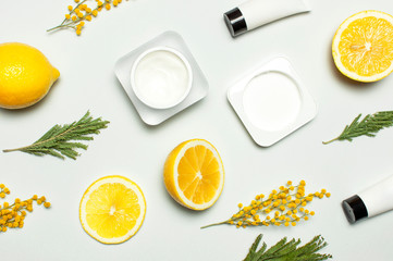 White cosmetic bottle containers with yellow Mimosa flowers and fresh lemon on gray background top view flat lay. Cosmetics SPA branding mock-up, Natural organic beauty product concept vitamin C