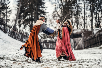 Fototapeta premium Battle of two knights in colored robes and hats near forest and wooden fortress. Knights fight in winter, in the snow
