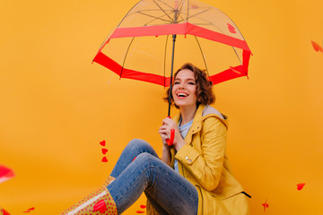 Magnificent young woman in jeans and yellow coat posing under parasol. Indoor photo of fashionable...