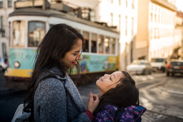 Latin mother and daughter looking affectionately outside on a Lisbon street, in the background there is a classic tram from Portugal