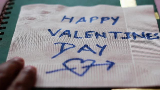 Moving a handwritten Happy Valentines Day message on a paper napkin over a notebook in a still frame shot.