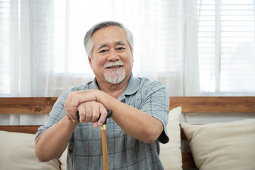 Portrait of senior old elderly asian man sit on coach hand hold help walking stick sit on sofa in house look at camera with happiness and healthy lifestyle.