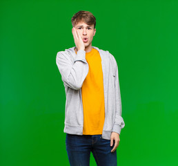 young blonde man feeling shocked and astonished holding face to hand in disbelief with mouth wide...
