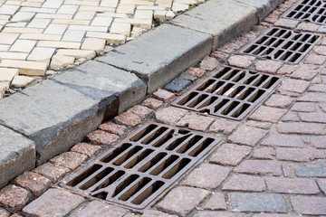 grille of the drainage system manhole on the paving road made of stone tiles by the curb, urban...