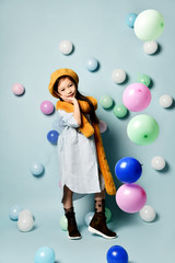 Fototapeta premium Little brunette asian girl in oversized shirt dress, brown beret and scarf, She posing with balloons on blue background.