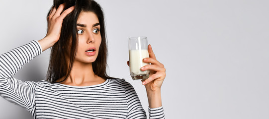 Excited young woman holding a glass of milk, not knowing whether to drink it or not milk. - obrazy, fototapety, plakaty