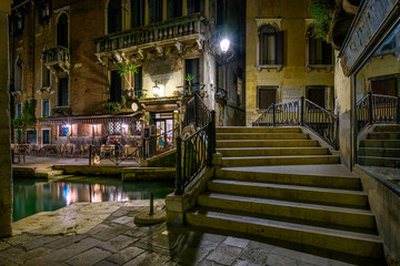 Narrow canal with bridge and tables of restaurant in Venice, Italy. Architecture and landmark of Venice. Night cozy cityscape of Venice.