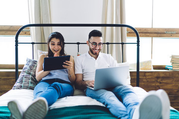 Cheerful couple entertained on gadgets in bedroom
