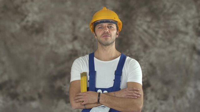 Studio portrait of serious handsome muscular young builder with coveralls, helmet, holding a folding rulerl ooking at camera and smiling. Portrait of worker about occupational safety. Shot in 6k.