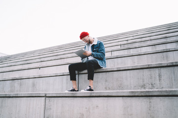 Happy fashionable male sitting on gray bench and using tablet