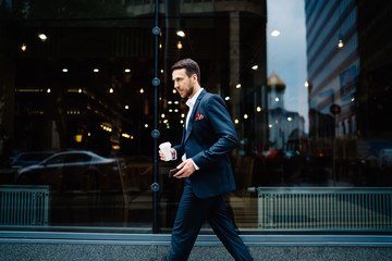 Tense businessman in stylish suit with smartphone and coffee to go walking in street in downtown