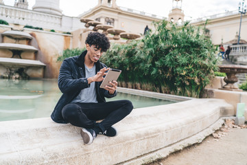 Young black man sitting on side of fountain choosing app on tablet