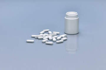 A pile of white pills lying in front of a small plastic white bottle , and several pills in it. Pills and bottles are white. Blue background