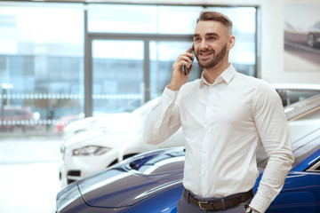Charming man talking on the phine at car dealership, looking excited after buying new auto