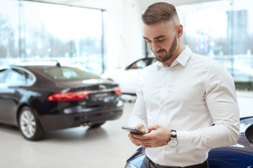 Handsome young man using smart phone at car dealership, copy space. Man texting on his phone, leaning on a car at auto salon