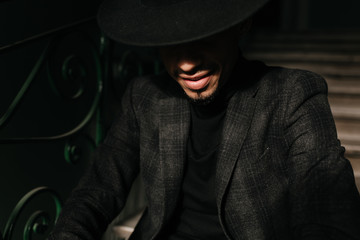 Obraz premium Close-up portrait of african man in formal wear posing in the dark. Refined male model in big hat sitting on stairs.