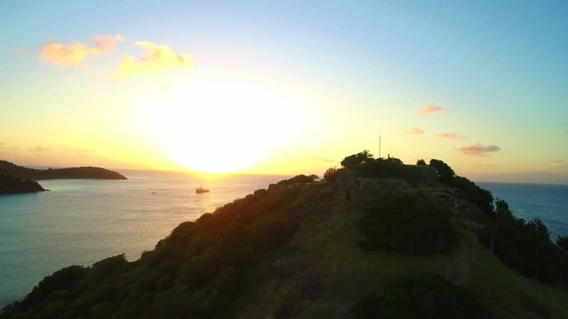 Beautiful cinematic drone shot of golden reflecting sunset, flying over an old historic fortress overlooking beautiful still gentle Caribbean sea with boat docked.