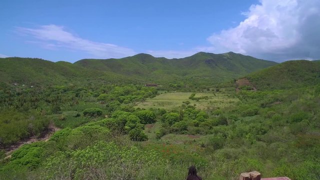 Reveal drone shot over abandoned house flying over black rasta man who's looking out over a valley of lush greenery and mountain range