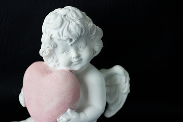 White angel with a pink heart in hands on a black background.