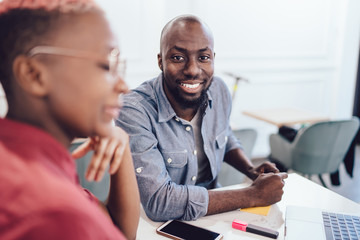 Pleased black man sitting at table with gadgets beside coworker