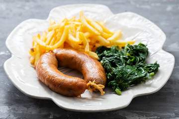 fried smoked sausage with potato chips and greens on beautiful plate on ceramic background