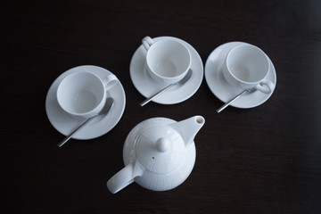 Light tea dishes are placed on a dark table. A white teapot and three mugs with saucers and spoons.