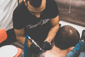 Fototapeta na wymiar Tattoo salon. The tattoo master is tattooing a man on his shoulder. Tattoo machine, safety and hygiene at work. Close-up, tinted, tattooist