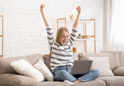 Happy blonde girl celebrating success, using laptop at home