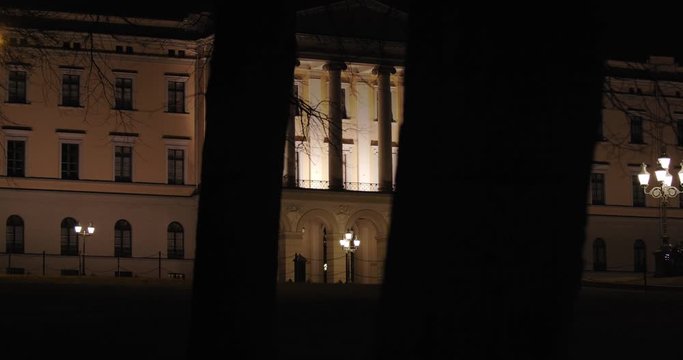 Slow motion close-up to wide 4K shot with parallax motion of dark trees moving in front of illuminated Norwegian Royal Palace on top of Karl Johan street, at night in Oslo Norway.