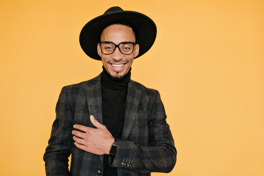 Happy Black Young Man Wears Elegant Dark Suit Posing With Pleased Smile. Indoor Photo Of Relaxed Male Model In Glasses Enjoying Photoshoot.